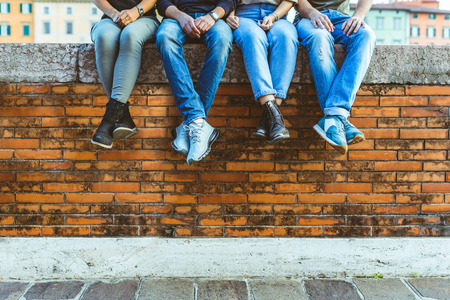Legs of four teenagers sitting on a bricks wall. Close up view on bottom part of the body, two girls...