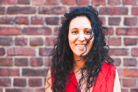 Smiling woman portrait against brick wall background. Happy girl with mixed race black and caucasian features and curly hair looking at cameraの写真素材