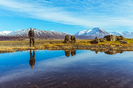 Man explorer in Iceland with his reflection on the water. Adult man walking and looking around. Majestic mountains on background, reflections on water on foreground. Travel and wanderlust conceptの写真素材