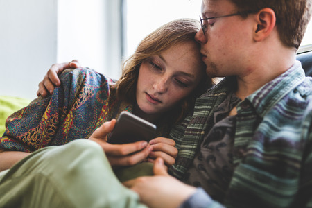 Romatic couple cuddling and embracing on the sofa. Woman holding a mobile phone and looking at it, man kissing her forehead in a romantic and candid moment.の写真素材