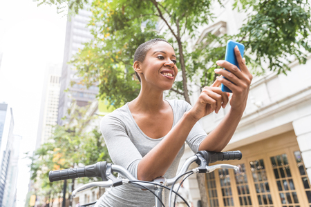 Black woman with bike typing on the phone in the city. Beautiful woman wearing a gray dress, holding her smartphone and moving around in the city in a eco friendly way. Lifestyle and relax conceptsの写真素材