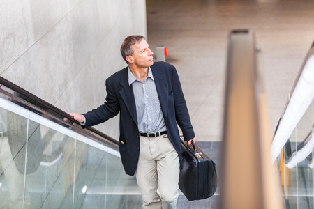 Adult businessman with suitcase on the escalator in the city. Caucasian man on his late fifties commuting in Berlin, walking out of a station. Business and city life conceptsの写真素材