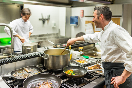 Chef and cook at work in a restaurant kitchen . Teamwork concept in working environment, two men cooking and wearing white uniform and black apron.の写真素材