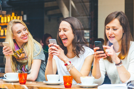 Three beautiful women in a cafe in Copenhagen enjoying coffee break together. Girls, best friends, laughing and smiling as they look at their smartphones. Lifestyle, friendship and happiness conceptsの写真素材