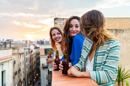 Beautiful women enjoying a beer on a rooftop in Barcelona. Three girls drinking a beer and having fun during a party at sunset on a rooftop with Gothic quarter on background in Barcelonaの写真素材