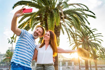 Adult couple dating and having fun in Barcelona at sunset. Man and woman together on a summer sunny day in Barcelona, taking a selfie with palm trees and sun on background. Love and lifestyle conceptsの写真素材