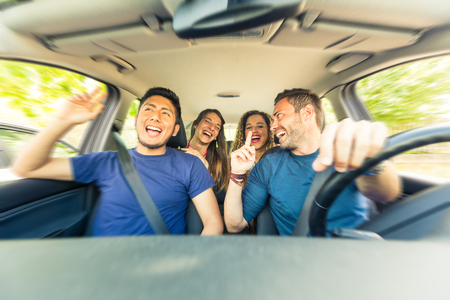 Friends inside the car singing during a road trip . Multicultural group of friends leaving for vacations. Two men sitting on the front and two women on the back singing, laughing and having fun