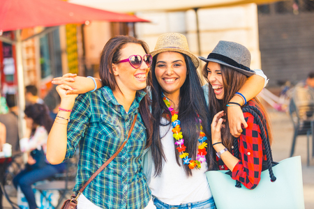 Three happy young women in the city, laughing, smiling and embracing. Girls friends mixed race group, summer shot with sunglasses, hats and accessories. Lifestyle, friendship and urban life concepts.の写真素材