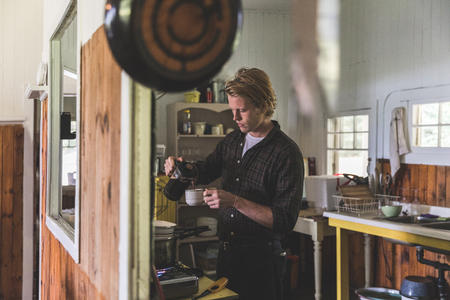 Man pouring coffee in the kitchen on a old wooden cottage. Handsome blond man wearing a checked shirt preparing breakfast and cooking. Lifestyle and travel conceptsの写真素材