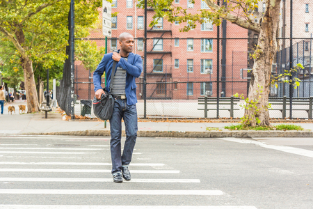 Young black man in New York crossing the street on the commute to work. Urban scene with playground and buildings on background. Lifestyle and millennials conceptsの写真素材