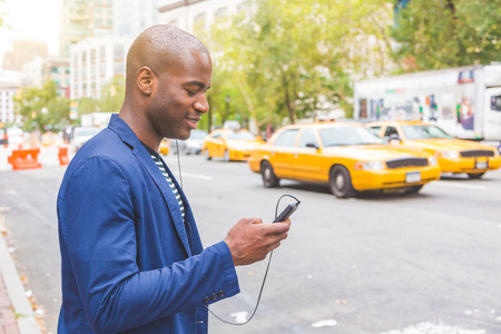 Young black man in New York looking at his phone on the commute to work. Street scene with yellow taxi cabs on background. Lifestyle and millennials conceptsの写真素材
