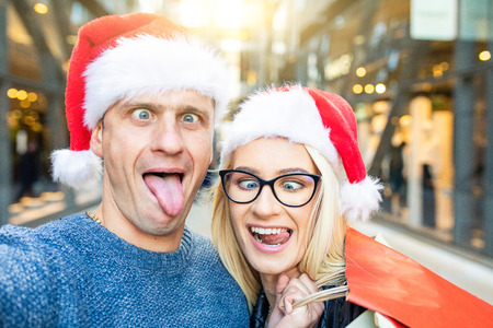 Couple shopping on Christmas time taking a funny selfie. Young caucasian couple, man and woman with Santa Hat and shopping bags making faces to the camera. Lifestyle and holidays.の写真素材