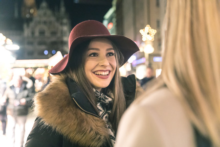 Happy women meeting and chatting at Christmas market in Munich. Brunette woman smiling at her blonde friend and looking at her. Christmas lights on background.の写真素材