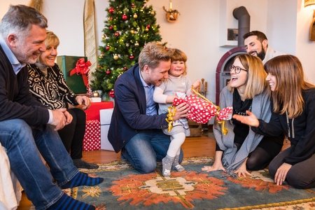 Happy little girl with family unboxing gift on Christmas day at home. Playful festive situation with blonde funny girl holding a present. Christmas and holidays conceptsの写真素材