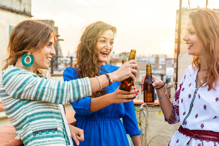 Happy women enjoying a beer on a rooftop in Barcelona - Three girls drinking a beer and having fun during a party at sunset on a rooftop with Gothic quarter on background in Barcelonaの写真素材