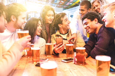 Happy millennial friends at pub drinking beer - Group of multiracial people having fun together at pub and looking at smartphone - Birthday party or after work meeting, happiness and teamwork concepts