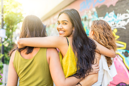 Beautiful woman embracing girl friends walking in the city - Urban portrait of a mixed race young woman looking back at camera and smiling - Diversity and lifestyle concepts in multicultural London cityの写真素材