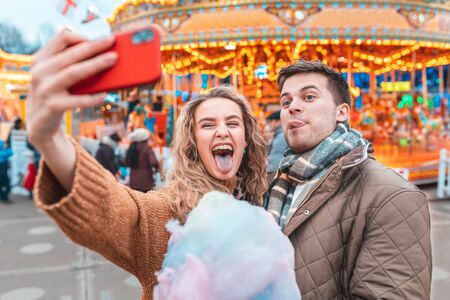 Couple having fun and taking a selfie at amusement park in London - Young man and woman making funny faces at camera and eating cotton candy at funfair - Happy lifestyle and love conceptsの写真素材