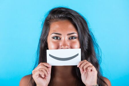 Happy smiling asian girl with emoticon smile - Happy young mixed race woman holding smiley emoji and looking at camera, blue plain background - happiness and successの写真素材