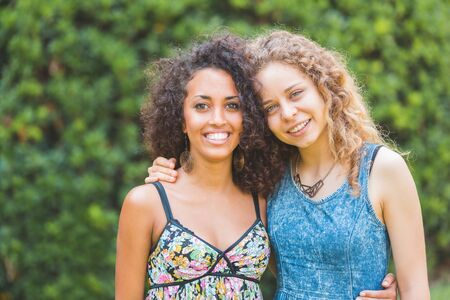 Multiracial happy young women portrait at park - Lovely female couple embracing and looking at camera - Mixed race black and caucasian curly girls sharing happinessの写真素材
