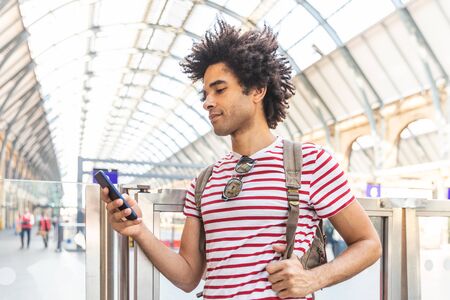 Happy man using the phone at train station in London - Mixed race young man with curly hair smiling and typing on the phone, waiting for train - Backpacker travel and lifestyleの写真素材