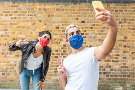 Happy couple taking a selfie with face mask on - Young man and woman taking a self portrait together and greeting the camera with hand gestures - lifestyle and happiness during coronavirurs pandemicの写真素材