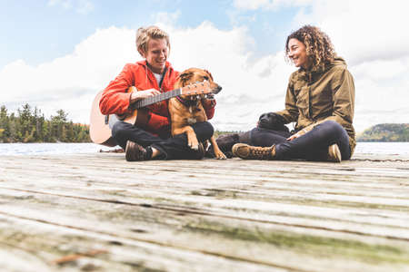 Happy young couple playing with their dog and guitar on a dock - Man and woman in love near a lake in Canada in autumn - love and lifestyle in the natureの写真素材