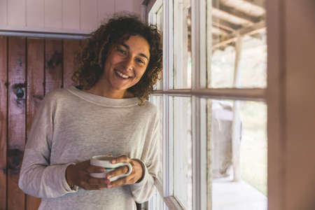 Woman smiling and looking at camera while holding a cup of coffee or tea - Beautiful and happy young woman with curly hair standing next to the window in a wooden cottageの写真素材