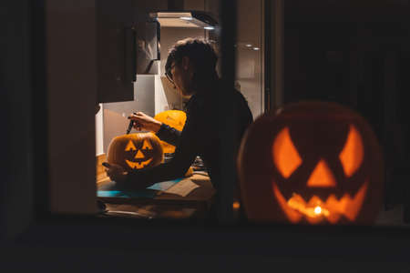 Woman carving pumpkin for Halloween night - View through the window of a young woman in the kitchen preparing pumpkins for the scary night - holidays, culture and lifestyle in UK and United States