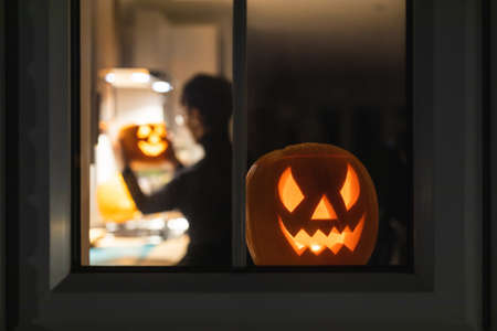 Woman carving pumpkin for Halloween night - View through the window of a young woman in the kitchen preparing pumpkins for the scary night - holidays, culture and lifestyle in UK and United Statesの写真素材