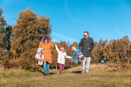 Happy family enjoying a day out together on a sunny autumn day - Mother and father holding and swinging their children, two cute kids, brother and sister, and having fun all togetherの写真素材