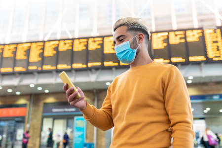 Man wearing face mask at train station in London - young man looking at his smartphone with departure arrivals board behind - health and travel concepts during coronavirus pandemicの写真素材