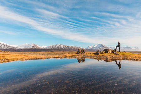 Explorer in Iceland with reflection on the water - Adult man standing on a rock looking around - Majestic mountains on background, reflections on water on foreground - Travel and wanderlust conceptsの写真素材