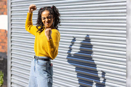 Happy black woman funny portrait showing muscles - Beautiful mixed race young woman wearing yellow top and curly hair playing with her shadow - happiness and lifestyle conceptsの写真素材