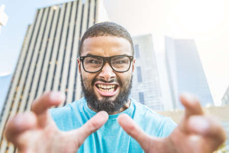 Black man shouting at camera - Angry young man wearing black eyeglasses looking at camera and raising hands - City buildings on backgroundの写真素材