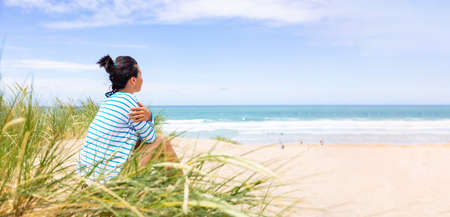Woman at seaside in Cornwall looking at beach and waves - Rear side view of a woman sitting on sand dunes with ocean on background on a sunny day - Travel and happiness conceptsの写真素材