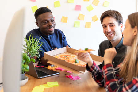 Happy employee people sharing pizza at startup office - Multiracial group of coworkers in small home office eating slices of pizza - Startup business or friends sharing a projectの写真素材