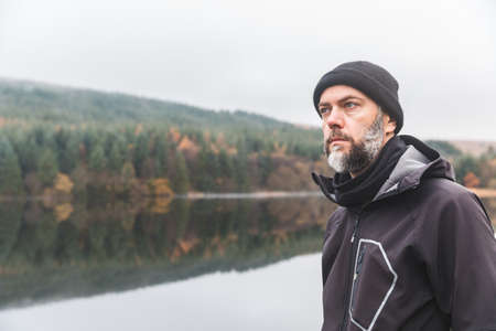 Man with beard portrait outdoor in autumn - Adult man wearing warm clothes and wool hat standing by a lake and looking away - Autumn style portraitの写真素材