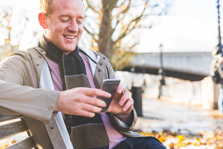 Happy smiling man portrait using phone in London - Young man sitting on a bench and typing on a smartphone, sharing life on social media and communicating with friendsの写真素材