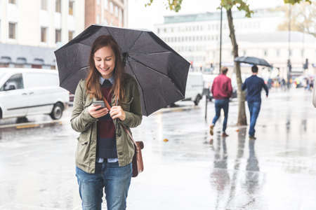 Happy woman using her phone on a rainy day in London - Young woman smiling, holding an umbrella and...