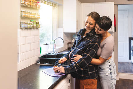 Authentic shot of happy married homosexual female gay couple expecting a baby and eating healthy apple together in the kitchen - lesbian couple at home enjoying life togetherの写真素材