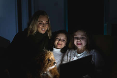 Authentic shot of two happy girls together with mum and dog playing with a digital tablet at home - Home scene of family having fun together while using technology device sitting on the sofaの写真素材