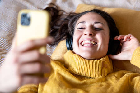 Authentic happy woman listening to music and singing lying on the bed - Beautiful smiling young woman wearing yellow top and holding her smart phone and headphonesの写真素材