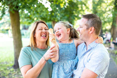 Authentic happy family having fun together at park with little girl playing with ice cream cone and...