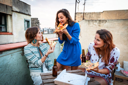 Happy women enjoying dinner party on a rooftop in Barcelona. Three girls drinking beer and eating pizza having fun during a party at sunset on a rooftop with Gothic quarter on background in Barcelonaの写真素材
