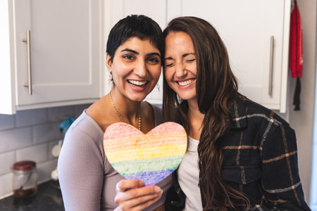 Authentic shot of happy lesbian couple holding a heart shaped rainbow flag and laughing - lesbian couple at home enjoying life togetherの写真素材
