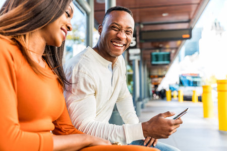Black couple waiting for the bus at station in Toronto - Man and woman sitting on a bench looking each other and laughing - Man using smartphoneの写真素材