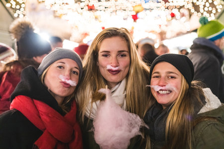 Girls eating cotton candy and making dummy moustache at Christmas market - Three happy young women enjoying time together and having funの写真素材