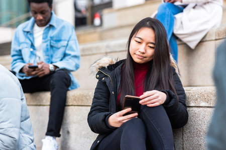 Multiracial millennial people using smart phones - Asian woman on foreground and holding a mobile smartphone, urban settings- College students or friends sitting and having a break at universityの写真素材