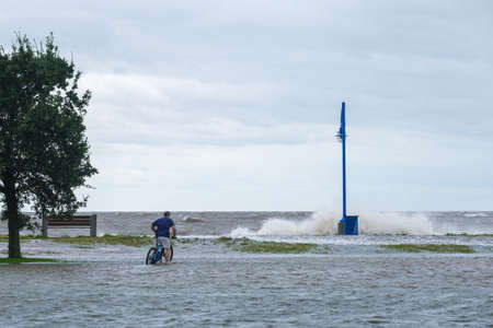 New Orleans, Louisiana/USA - 9/15/2020: Man riding bicycle in flooded street with wave crashing in background during Hurricane Sally storm surge on Lake Pontchartrainのeditorial素材